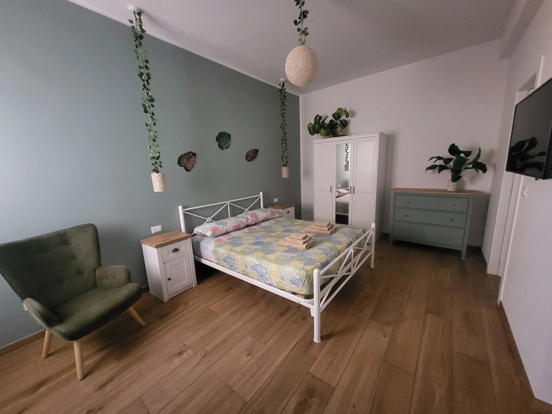 La camera vista dalla poltrona: parete verde salvia con piante pensili, armadio e cassettiera